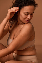 Portrait of a dark-haired curly woman with skin flaws, cellulite and stretch marks posing in beige underwear, looking down against colored background with copy space. Body positivity concept - obrazy, fototapety, plakaty
