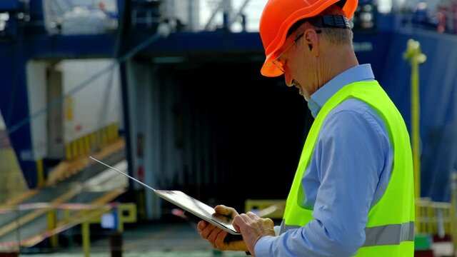Cargo ship loading in dock. Safety engineer with glasses inputs data of freight loading on tablet computer against open vessel hold in dockyard