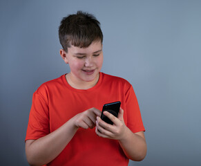 A smiling young man in a red T-shirt looking at his smartphone while exchanging text messages. A teenager with a phone, isolated on a gray background.