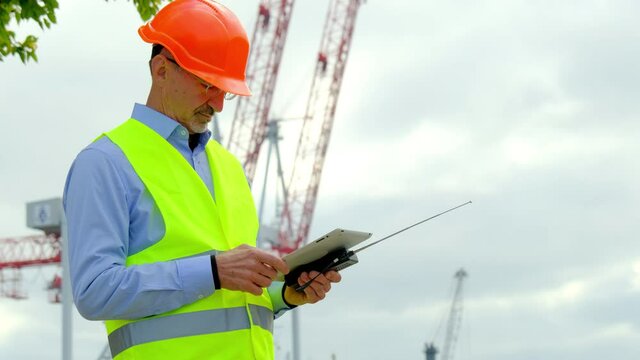Port worker at sea ships construction. Happy employee with tablet and walkie-talkie walks after finished cargo checkup against cranes in dock