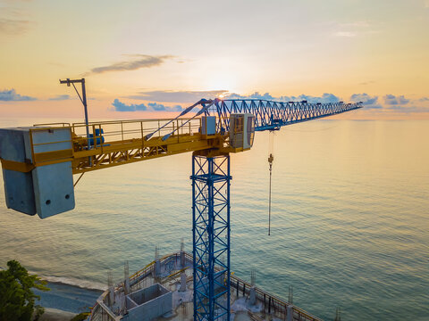 Close-up view from a drone of a construction tower crane against the background of a fantastic, beautiful sunset on the sea with a copy of the space