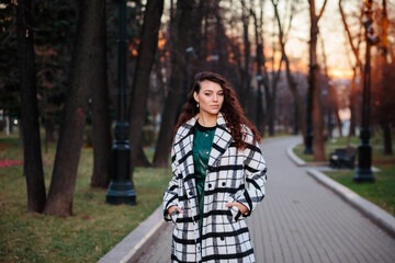 Glamorous woman in checkered coat and green leather dress is spending time outdoors in autumn park.
