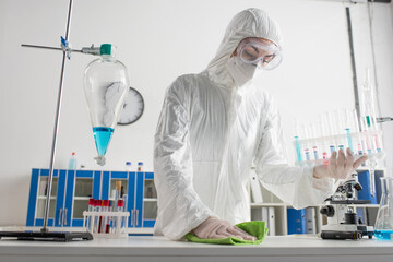 doctor in protective suit holding test tubes while wiping desk in laboratory.