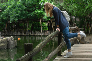 A girl bent over to an artificial pond in a public park and looks into the water from a wooden bridge. she has a white backpack on her shoulders. Travel and excursions alone, enjoying nature.