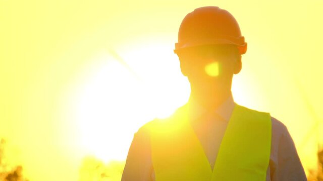 Worker in hardhat at sunset. Foreman in uniform walks at construction site of rural substation illuminated by bright sunlight closeup