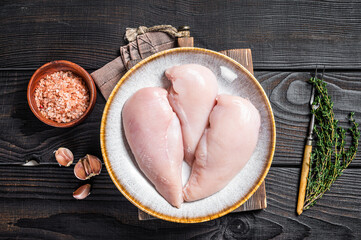 Chicken breast fillet raw meat in plate with herbs. Black Wooden background. Top view