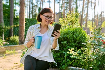 Fototapeta premium Middle-aged woman having rest in spring garden with cup of tea and smartphone in hands