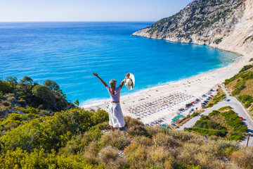 Happy woman standing on top of a rock, raising hands with an exciting feeling of freedom, looking at Myrtos Beach. Cephalonia island, Greece