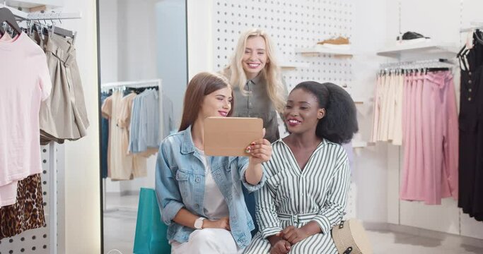 Three happy stylish women are sitting in mall and chatting with friends on video call using tablet, bragging about successful shopping.