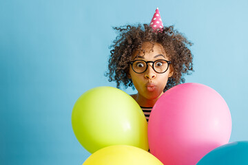 Young lady in birthday cone hat pouting lips and making funny face while holding colorful balloons....