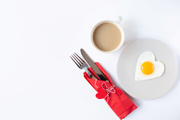 A cup of coffee, a plate with a fried egg in the form of a heart and cutlery in a red napkin with decorative hearts are on a white background. Valentine's Day breakfast theme. Free space for text.