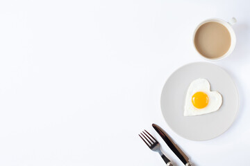 A cup of coffee, a plate with a fried egg in the form of a heart and cutlery are on a white background. Valentine's Day breakfast theme. Free space for text.