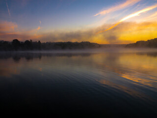 Sunrise over the foggy lake with the reflection of sun. Mist on the water, forest silhouettes and the rays of the rising sun. Beautiful morning landscape with sunrise over river.