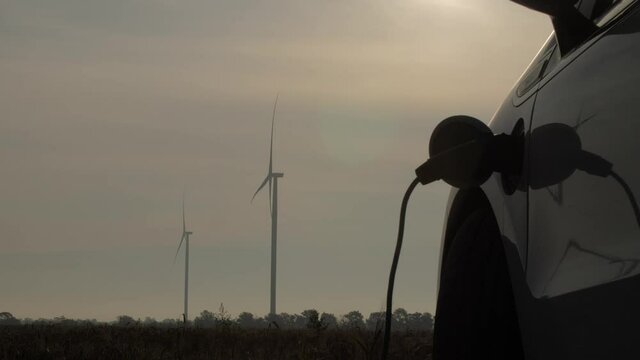 Windmills generate energy in field on cloudy weather. White electric automobile charged by alternative power against operating turbines close view