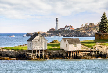 Old Fishing shacks per on a rocky shore of Fisherman’s Point with the Portland Head Light, a lighthouse along the Atlantic coast of Down East Maine in America’s New England region. - 472508995