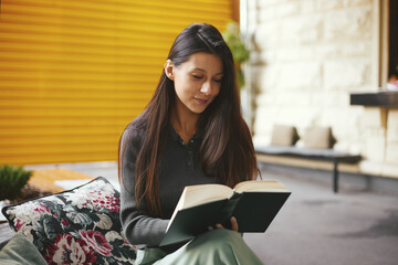 Portrait of adorable young woman reading book while sitting at coffee shop with cup with hot coffee