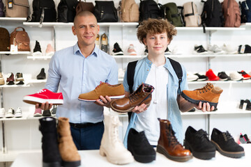 Portrait of happy adult man and teenage boy standing in shoe store holding loafers and boots in hands excited by large assortment