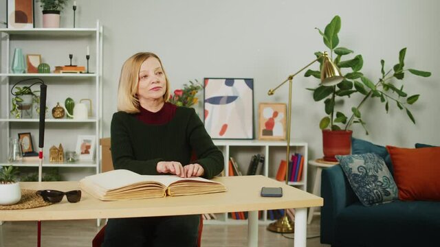 Blind woman reading braille book using fingers, sitting in living room, poorly seeing female person learning to read, home education for people with disabilities, touching letters on sheet of paper.
