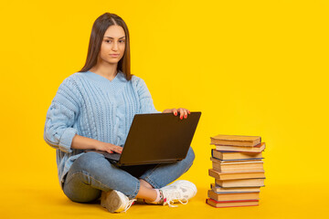 Young girl is preparing for exam. Stack of books in foreground. Student sits with laptop on her lap. Woman student is engaged in self-education. Preparing for university exam. Girl on yellow