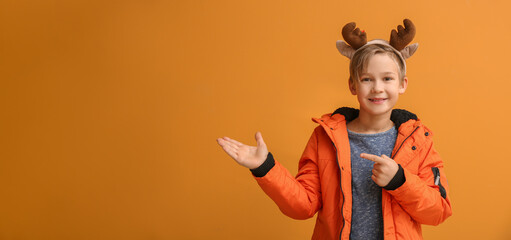 Cute little boy with deer horns and winter clothes showing something on color background with space for text