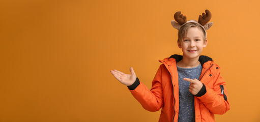 Cute little boy with deer horns and winter clothes showing something on color background with space...