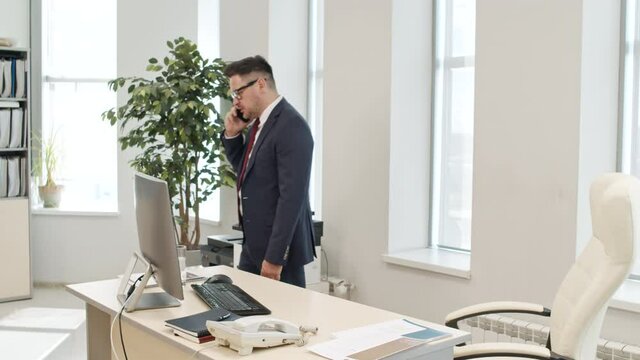 Handheld medium shot of angry male executive in suit and glasses pacing in his office and having animated conversation on mobile phone