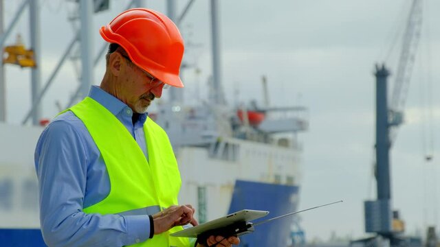 Sea industry port ship safety. Senior taskmaster in helmet checks drawings on tablet and looks at large repairing vessel in dock closeup