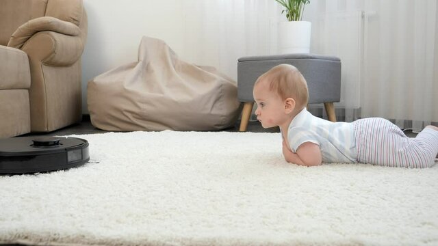 Robot vacuum cleaner rides to cute baby boy lying on carpet in living room. Concept of hygiene, household gadgets and robots at modern life.