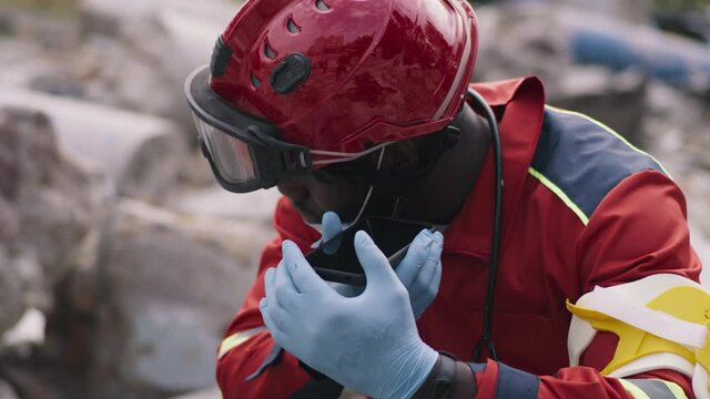 African American paramedic in red uniform listening audio message on smartphone while sitting on ruins of destroyed building after disaster