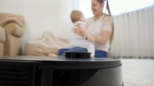 Robot vacuum cleaner working and doing clenup while mother playing with her child. Concept of hygiene, household gadgets and robots at modern life.