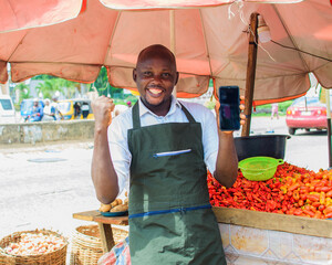 A happy African male trader with a smart phone, standing beside his stall of tomatoes and pepper in...
