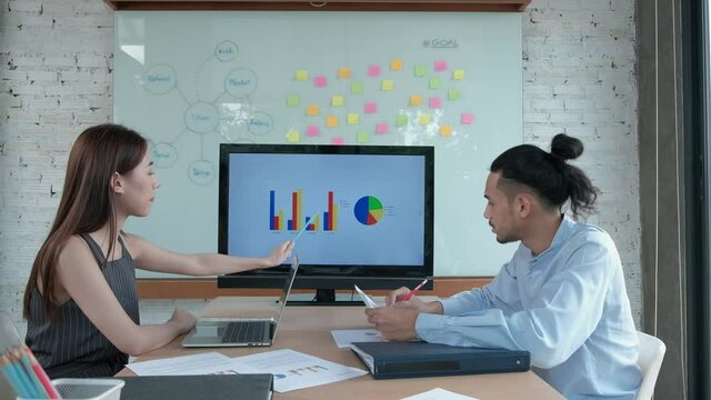 Two young and smart Asian coworkers are brainstorming at office. Female colleague presents business chart to male partner using a laptop in a meeting room, discussing the company's finances project.