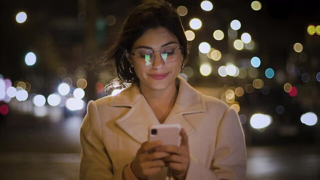 Lateral Dolly Attractive and Cheerful Young Woman Using Smartphone in Crowded Street. Texting on Mobile Phone at Night with Traffic.