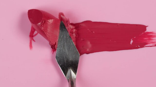 Smearing red lipstick with spatula close-up. Lip gloss on pink background. Professional makeup and decorative cosmetic, beauty products concept.