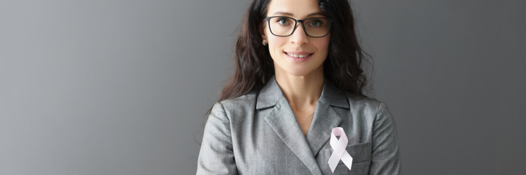Portrait of smiling woman in gray suit on her chest with pink ribbon symbol of fight against breast cancer - Powered by Adobe