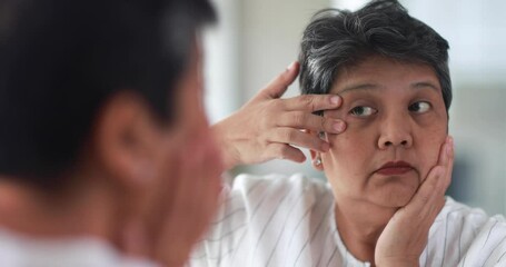 Old Asian woman feels stressed and serious face when watching wrinkle and aging sign around eyes and face in mirror. Elderly lady worrying about beauty. Concept of skin problems in senior people. - Powered by Adobe