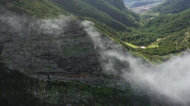 Epic and dramatic cliff wall with thin clouds moving around with a few small houses behind next to green grass fields and lush forest. Extreme and massive nature.
