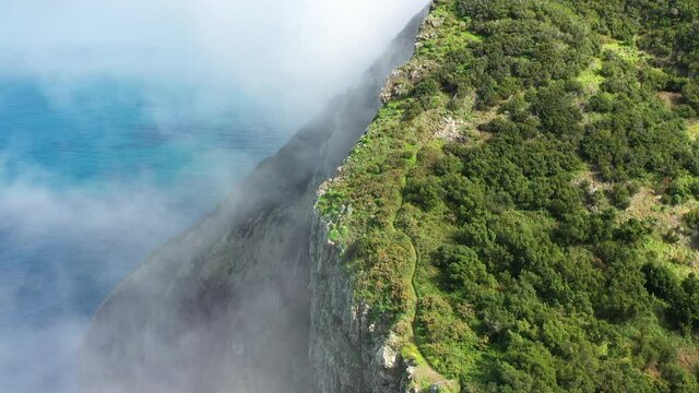 Thin clouds are moving slowly over the cliff on the coast of Madeira island with the turquoise and deep blue water below and green lush nature on the top with a small trail leading up the mountain.