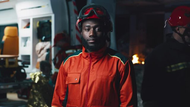 Black man in paramedic uniform looking at camera while standing near ambulance van and colleagues during rescue mission at night