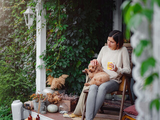 Young asian woman resting outdoors with cup of coffee on porch of country house, chilling outside with poodle dog . autumn lifestyle, leisure free time concept. Copy space