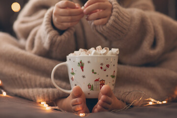 Child bare feet and hands hold cup hot cocoa marshmallow close up garland lights.flaffy fuzzy warm...