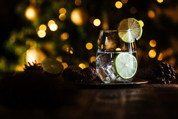 Gin tonic in cocktail glass with ice, and lime on a dark rustic board in Christmas decoration with Christmas tree and lights in blurry in background