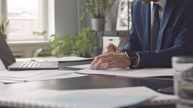 Low angle of cropped businessman wearing blue suit sitting at desk in office, taking notes with pen on document and using computer at daytime