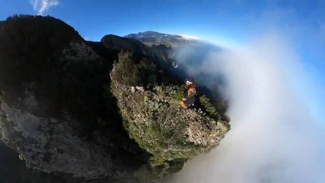 A young, fit and strong man is climbing and running alone, holding a selfie stick with 360 camera on the edge of Espigao Amorelo mountain in Maderia during a hot, sunny summer day with clear blue sky.