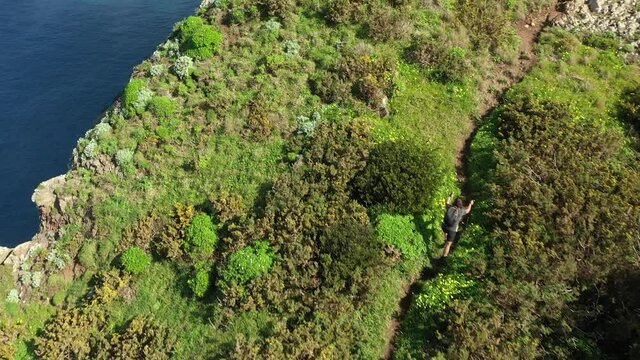 Hiker walking up a trail between lush and green grass, bushes and trees right next to the steep cliff down to the deep blue ocean on Madeira island in the summer sunshine.