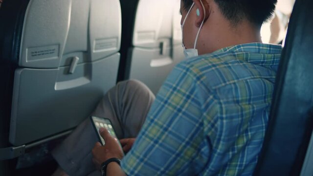 A young man of Asian appearance makes a flight on an airplane, a teenager uses a phone on an airplane, scrolls through pictures, messages, opens an application.