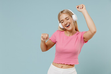 Young smiling happy cheerful caucasian blonde woman 20s wearing casual pink t-shirt headphones listen to music dance isolated on plain pastel light blue background studio. People lifestyle concept