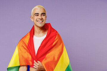 Young smiling hispanic latin gay man 20s with make up wrapped in rainbow striped flag look aside isolated on plain pastel purple background studio portrait. People lifestyle fashion lgbtq concept