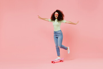 Full size body length happy fun excited bright young curly latin woman 20s wear casual clothes sunglasses skate on board spreading hands isolated on plain pastel light pink background studio portrait