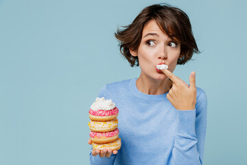 Young minded woman 20s wearing casual sweater look camera hold donuts dessert lick finger with cream isolated on plain pastel light blue background studio People lifestyle junk unhealthy food concept
